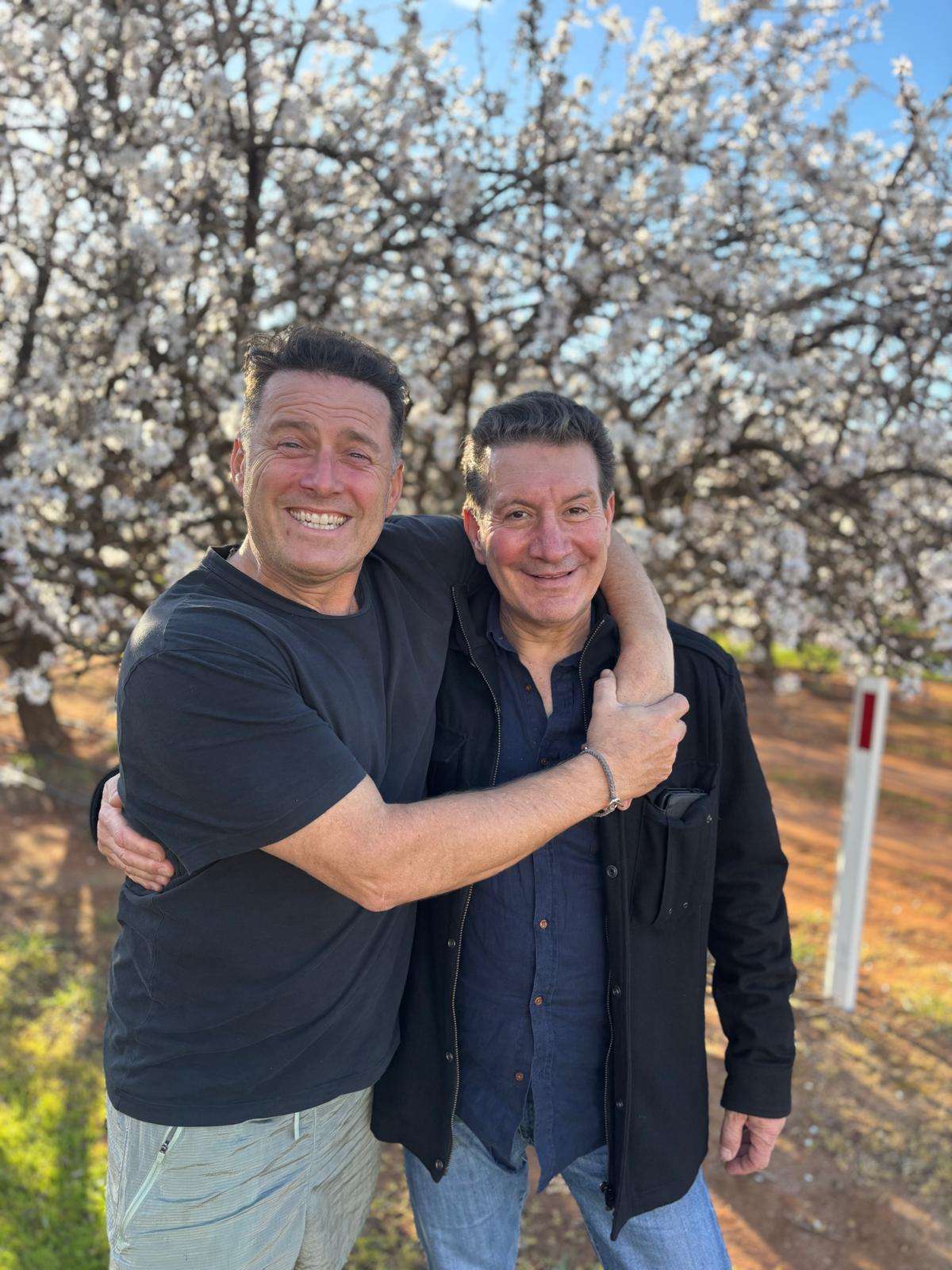 Roy Spagnolo shares a warm embrace with longtime friend and television presenter Karl Stefanovic beneath blossoming trees, smiling together during an outdoor gathering.