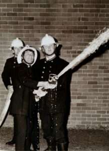 A black-and-white photograph from the late 1980s showing firefighter Roy Griffith during a training exercise, operating a high-pressure fire hose with fellow firefighters standing behind him.