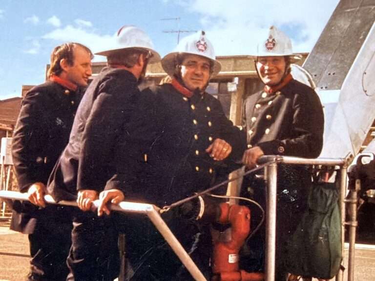 A late-1980s photograph of firefighter Roy Griffith standing on the far right with fellow crew members beside a fire service cherry picker, wearing full firefighting uniforms and helmets.